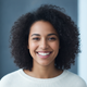 A happy young woman with dark, curly hair smiles genuinely at the camera, conveying trust and satisfaction.